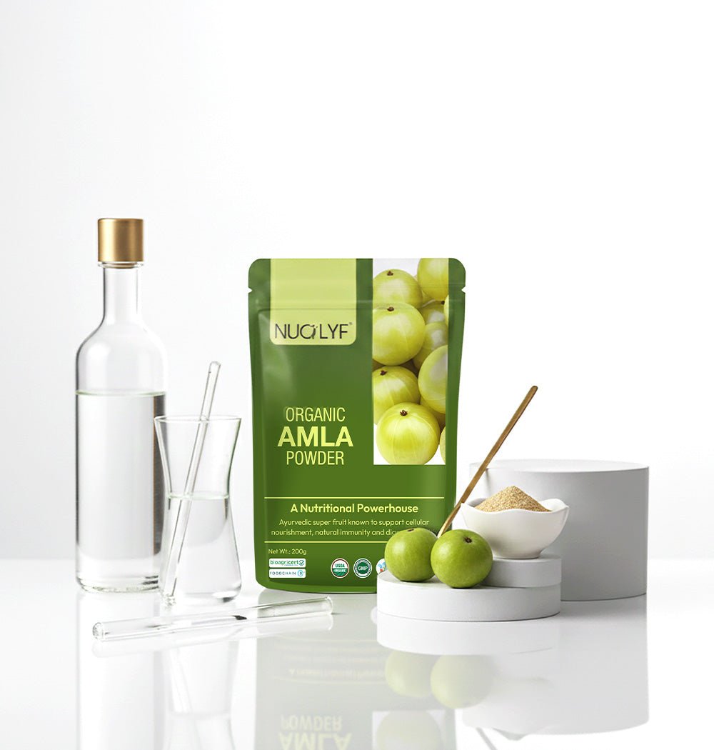 Organic amla powder pouch with fresh amla fruit, glassware, and powder in a bowl on a white surface