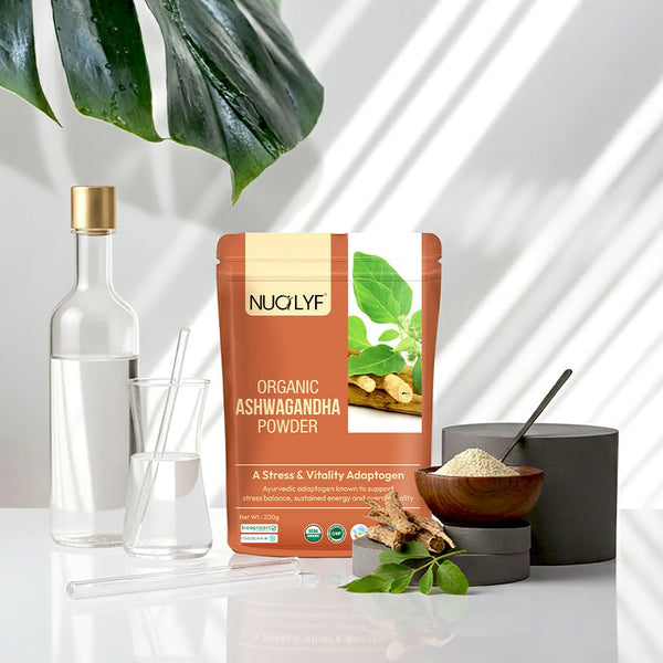 Organic ashwagandha powder pouch with glassware, leaves, and ashwagandha root on white table.