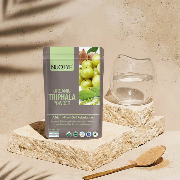 Organic Triphala powder pouch with fresh ingredients, glass of water, and wooden spoon on stone blocks